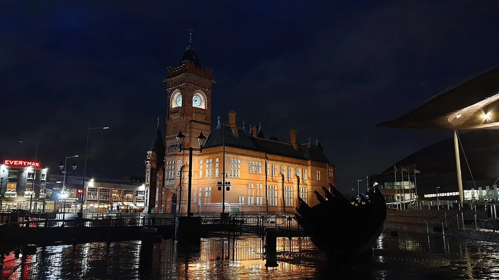 The Pierhead Building's red brick and clock tower lit up at night on the Cardiff Bay waterfront, with the Senedd alongside