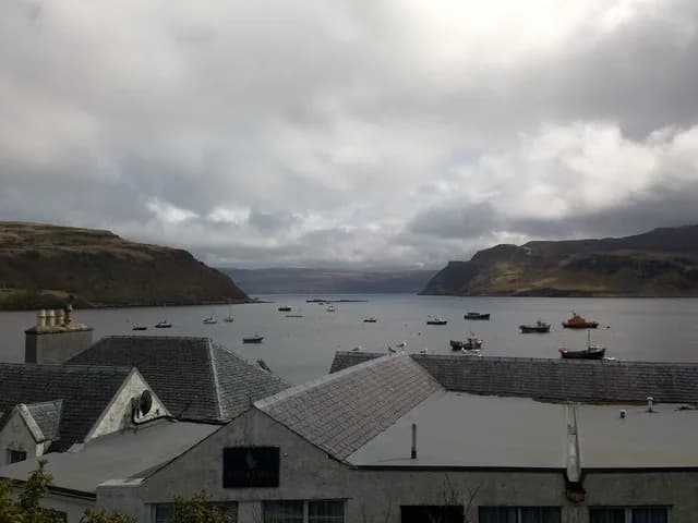 Moored boats in Portree harbour under grey cloud, seen over slate rooftops, with the headlands at the mouth of the bay beyond