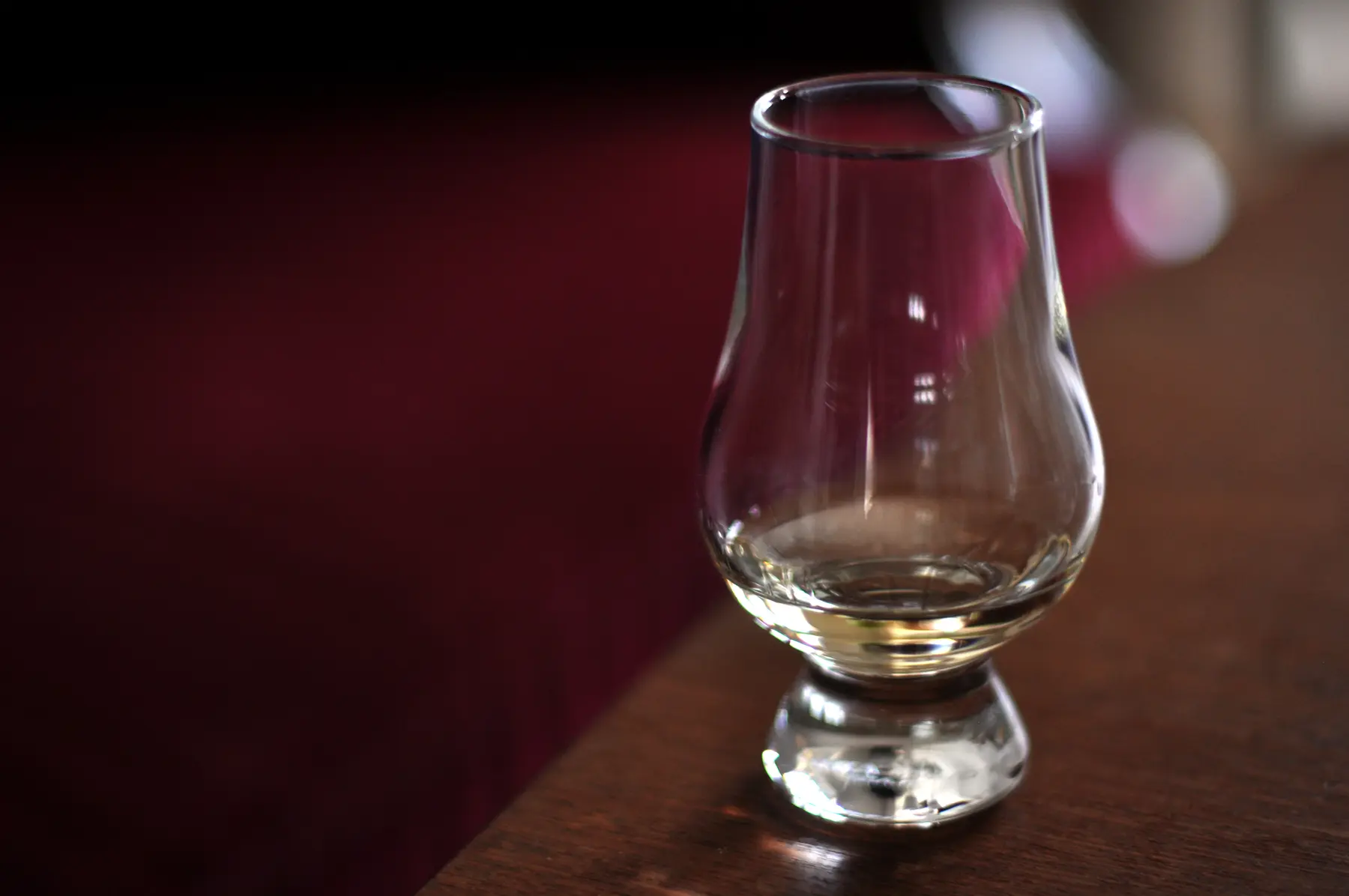 A Glencairn nosing glass holding a small measure of whisky, on a dark wood table against a deep red banquette