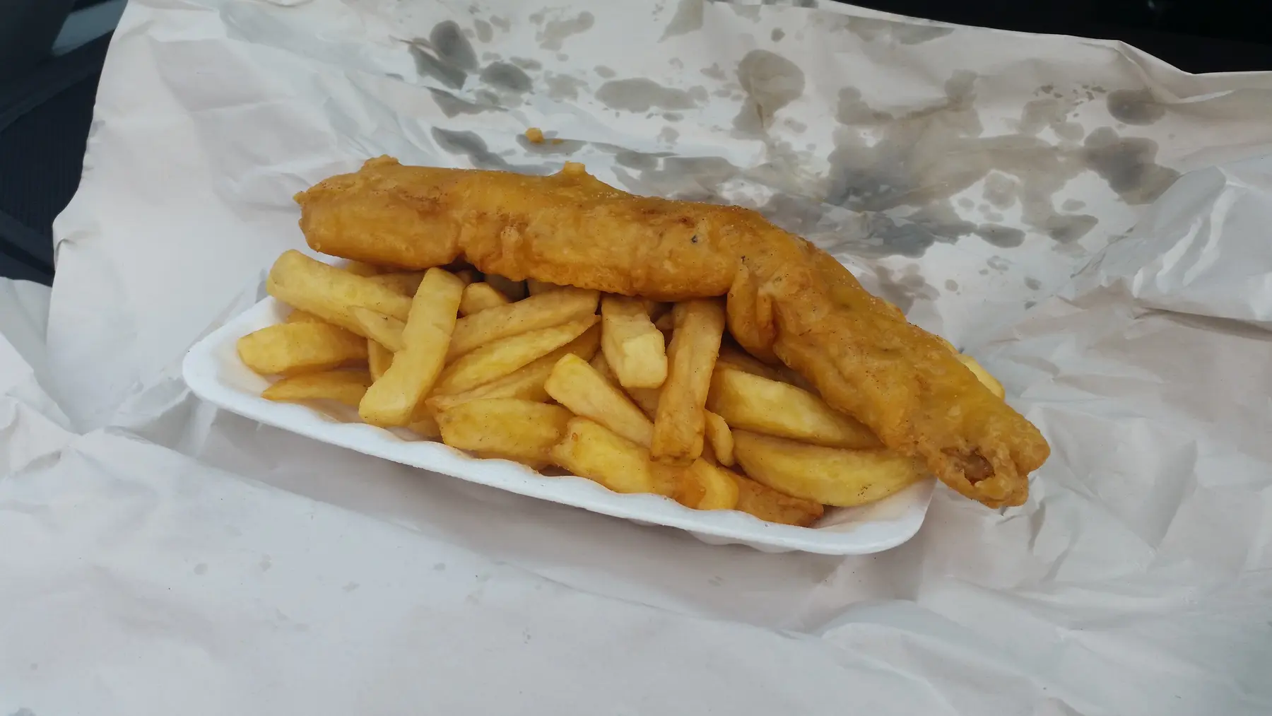 A portion of battered fish and thick-cut chips in an open tray on wrapping paper
