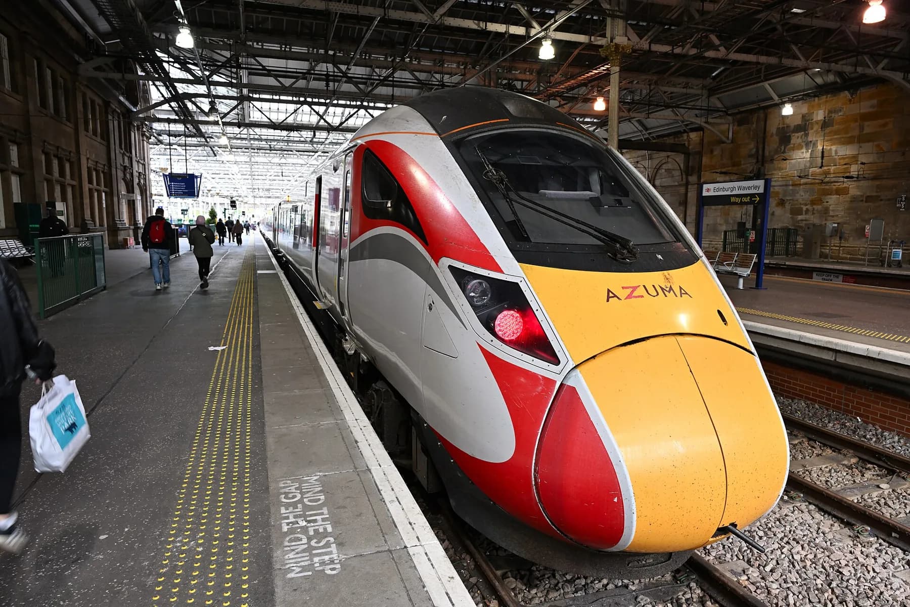 An LNER Azuma waiting at a platform at Edinburgh Waverley station, passengers walking along the platform under the station's iron roof