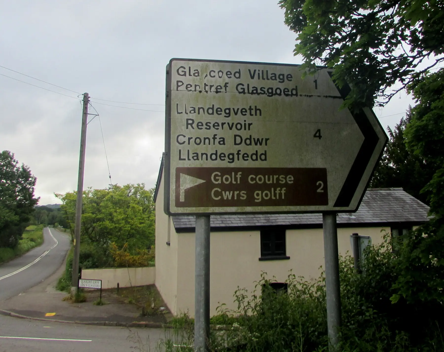 A weathered bilingual road sign on a rural lane at Monkswood, Monmouthshire, giving every destination in both English and Welsh, including Glascoed Village and Pentref Glasgoed