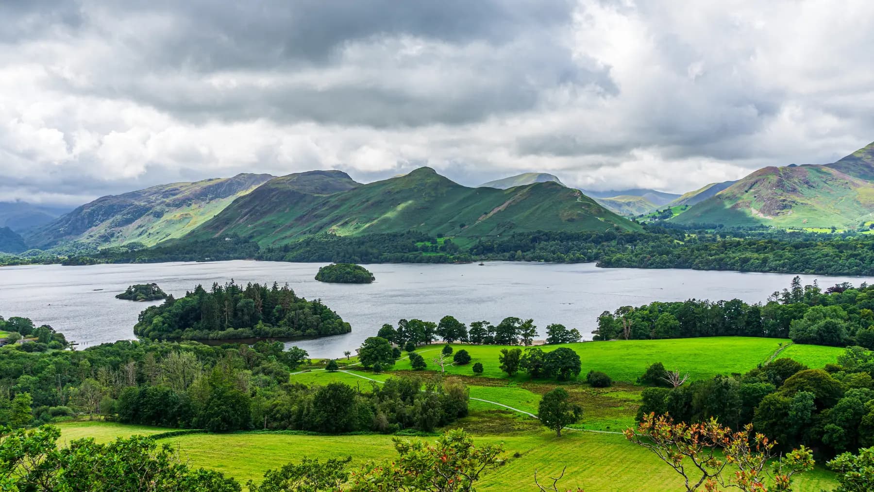 Derwent Water seen from Keswick in the Lake District National Park, with wooded islands on the water and a ridge of green fells rising beyond under heavy cloud