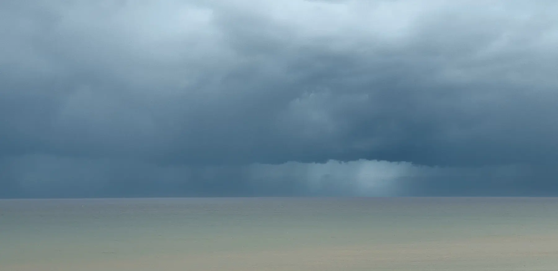 A bank of dark rain cloud over a flat grey sea, with a shower visibly falling on the horizon